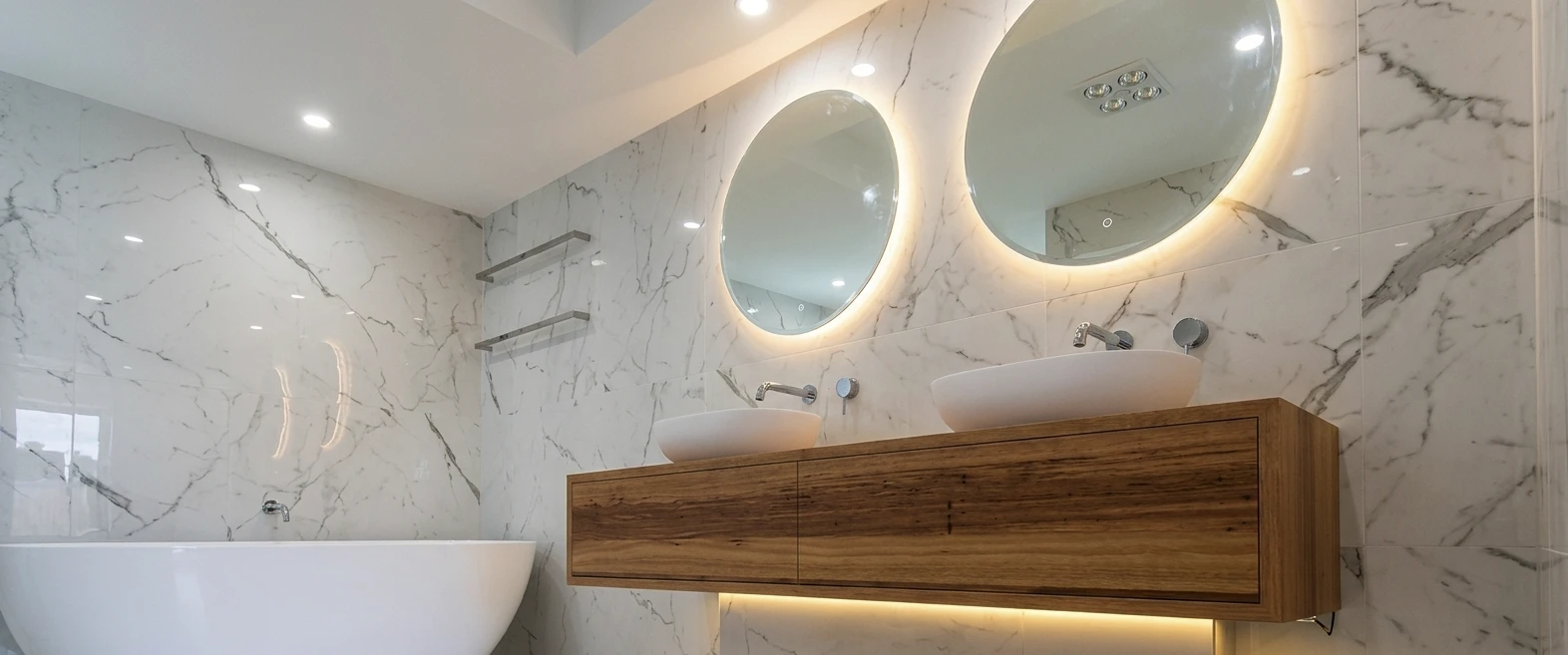 Bathroom with floor-to-ceiling white marble-look tiles, twin backlit round mirrors and a timber floating vanity