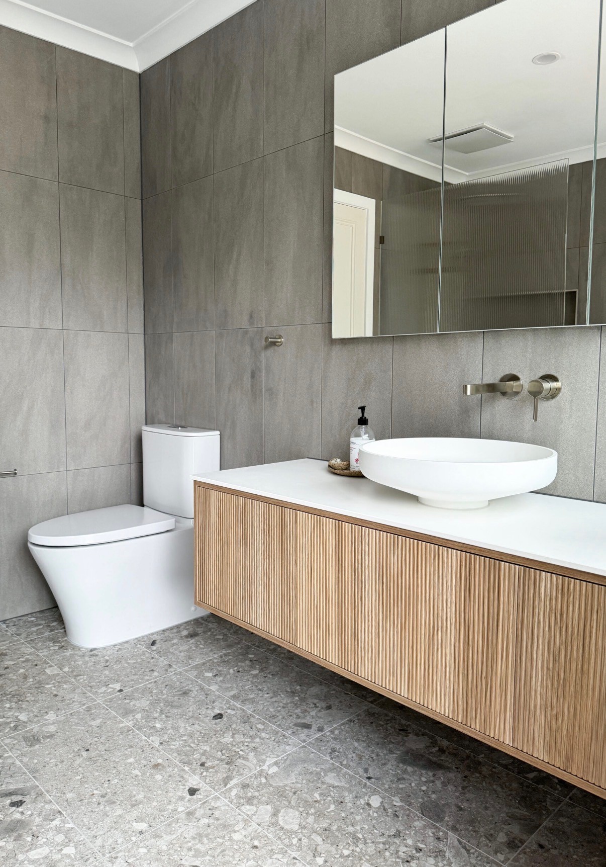 Bathroom with large-format grey wall tiles, a terrazzo-look floor and a fluted timber floating vanity with a white vessel basin
